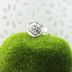 May include: Silver ring featuring a round, engraved image of a baby's face. The ring is displayed on a bed of green moss.