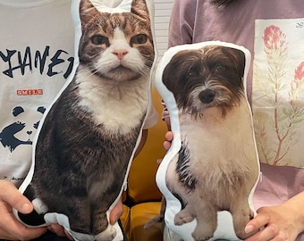 Può includere: Due cuscini personalizzati per animali domestici, uno con un gatto tabby marrone e l'altro con un cane bianco e nero. Entrambi i cuscini sono bianchi con un tessuto morbido e soffice.
