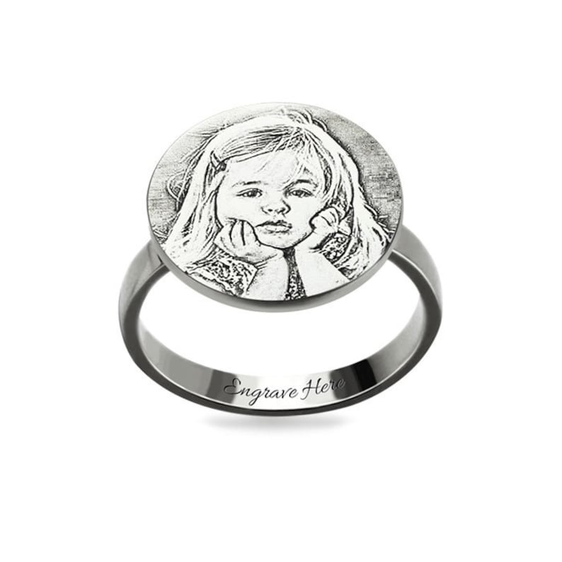 May include: A silver ring featuring a round, engraved photograph of a young girl. The ring has the words "Engrave Here" inscribed on the band.