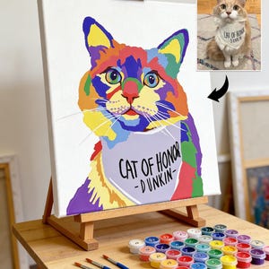 May include: A colorful cat portrait on canvas, displayed on a wooden easel. The artwork features a vibrant, multi-colored cat with the text "CAT OF HONOR - DUNKIN" on a bib. Paint pots and brushes are arranged below.