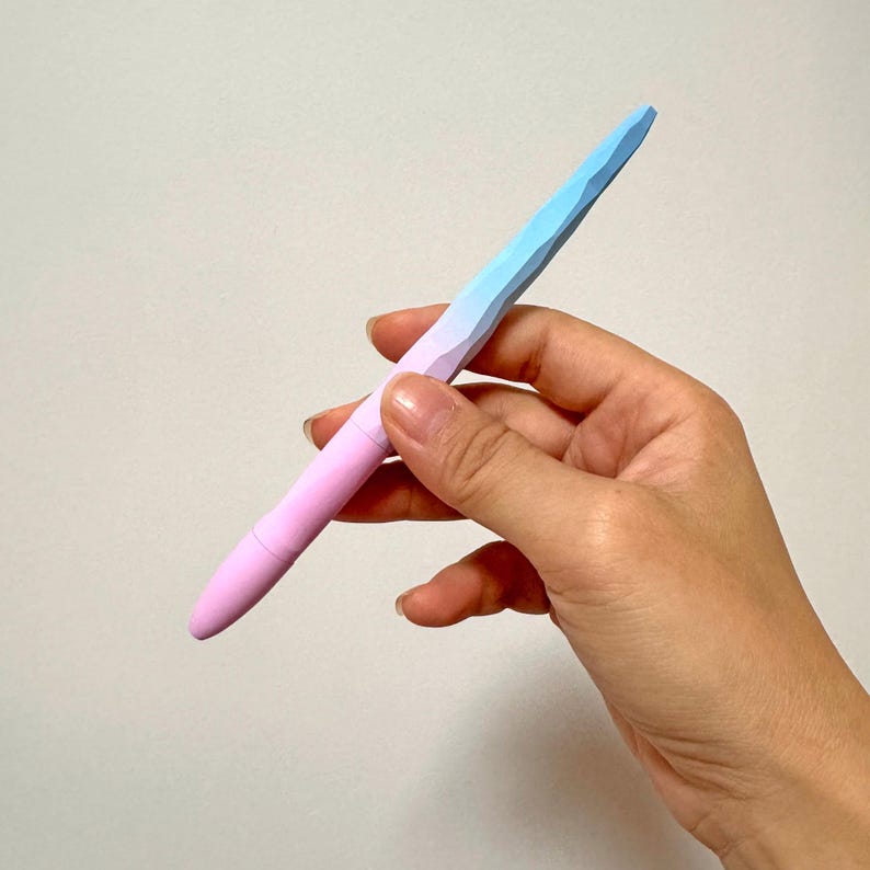 May include: A pastel-colored pen with a faceted design. The pen transitions from pink to blue, held against a neutral background. The pen's design is geometric, with a smooth, rounded base.