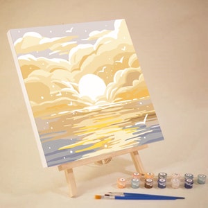 May include: A paint by numbers kit featuring a sunset over the ocean. The canvas is on an easel and the paint pots are arranged in a row below. The colors in the painting are yellow, orange, blue, and white.