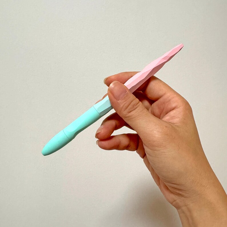 May include: A pastel-colored pen with a faceted design. The pen features a light blue barrel and a pink tip. The pen is held in a hand against a plain background. The pen's design is geometric and modern.
