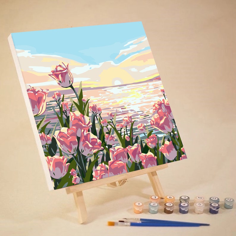 May include: A paint by numbers canvas depicting a sunset over a body of water with pink tulips in the foreground. The canvas is on an easel and there are paint pots with numbers on them.