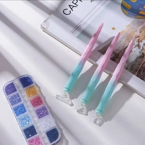 May include: A collection of diamond painting tools and supplies. The image shows four pink and blue gradient pens with clear tips, a clear plastic organizer filled with colorful diamond beads, and a magazine.