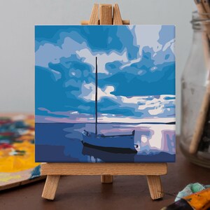 May include: A painting on canvas depicting a sailboat on a calm body of water with a blue sky and white clouds. The painting is displayed on a small wooden easel.