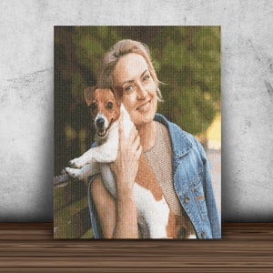 May include: A woman in a blue denim jacket is holding a small white and brown dog in her arms. The dog is looking at the camera and the woman is smiling. The background is a blurred green and brown.