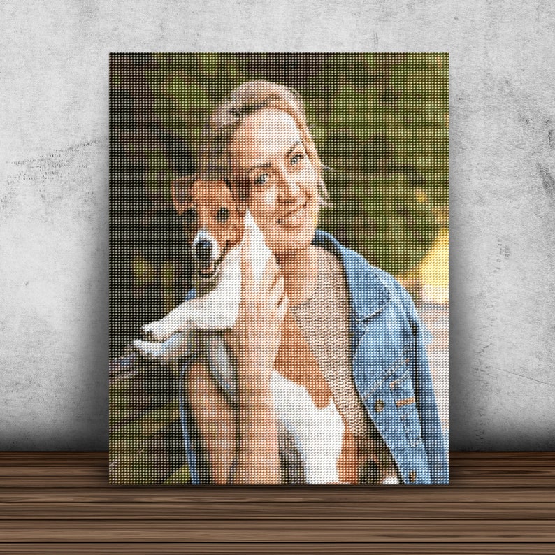 May include: A woman in a blue denim jacket is holding a small white and brown dog in her arms. The dog is looking at the camera and the woman is smiling. The background is a blurred green and brown.