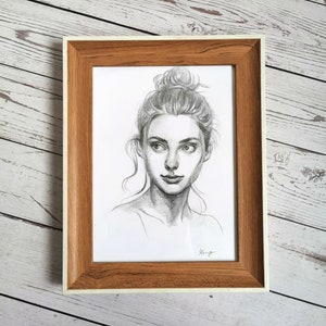 May include: A black and white pencil drawing of a woman with her hair pulled back in a bun. She is looking off to the side and has a soft expression on her face. The drawing is framed in a light brown wood frame with a white mat.