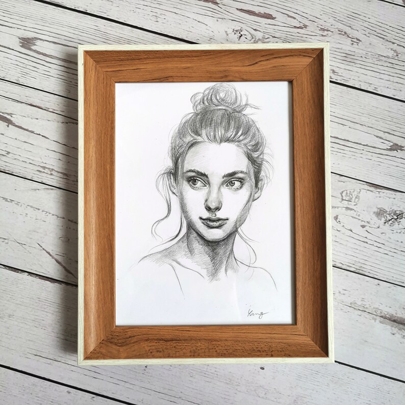 May include: A black and white pencil drawing of a woman with her hair pulled back in a bun. She is looking off to the side and has a soft expression on her face. The drawing is framed in a light brown wood frame with a white mat.