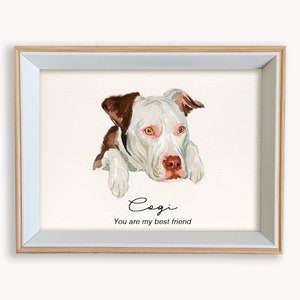 May include: A watercolor painting of a white and brown dog with brown eyes. The dog is looking at the viewer and has its head resting on its paws. The painting is framed in a light wood frame and has the text "Cogi You are my best friend" written below the dog.
