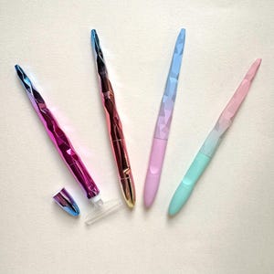 May include: Four colorful nail files with faceted, iridescent handles. The files are in shades of pink, purple, blue, and gold. One file has a detached cap and a clear plastic base. The files are arranged on a light-colored surface.