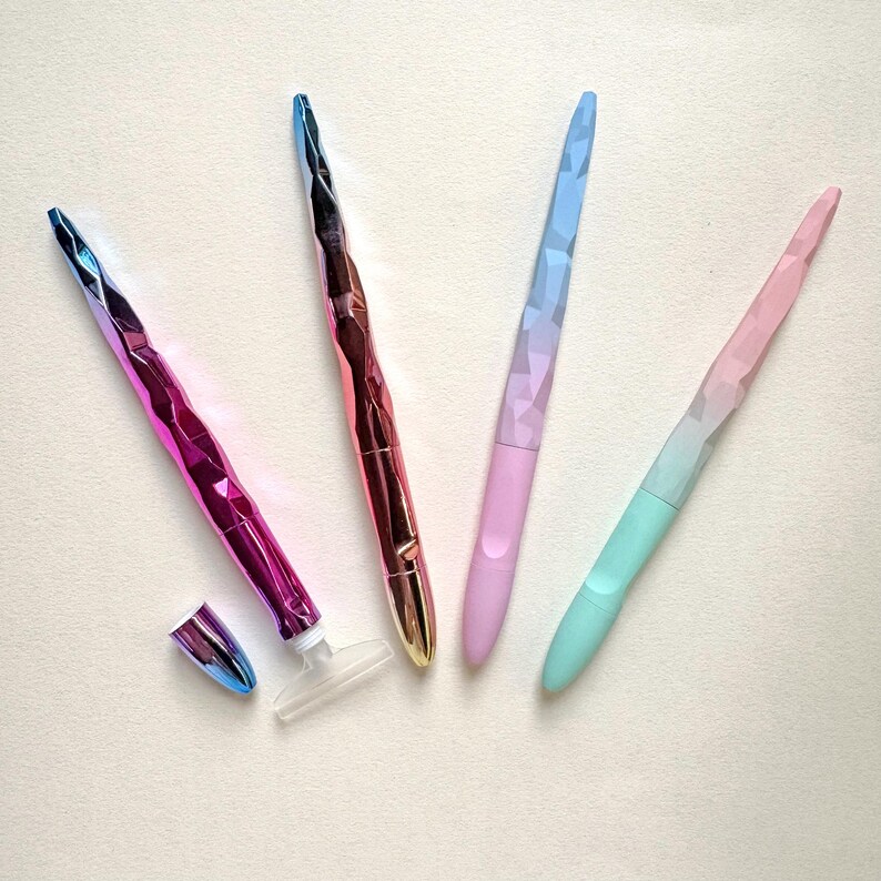 May include: Four colorful nail files with faceted, iridescent handles. The files are in shades of pink, purple, blue, and gold. One file has a detached cap and a clear plastic base. The files are arranged on a light-colored surface.