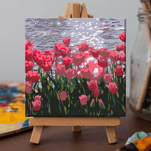 May include: A painting on canvas depicting a field of pink tulips with a blue water background. The painting is displayed on a small wooden easel.