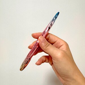 May include: A metallic pen with a faceted design. The pen transitions in color from gold to pink to blue. The pen is held in a hand against a white background. The pen's design is geometric and modern.