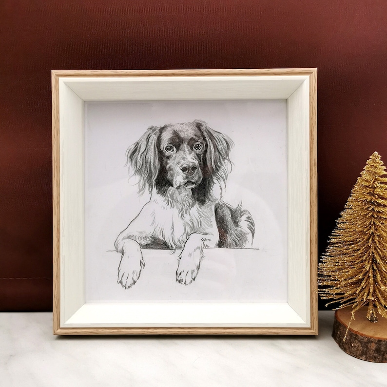 May include: A framed pencil sketch of a dog with brown and white fur, resting its paws on a ledge. The frame is light wood with a white mat. A small gold glitter Christmas tree sits to the right.