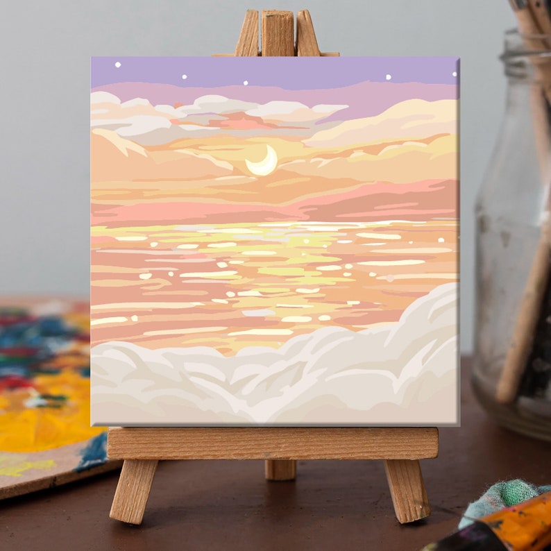 May include: A painting on a small easel depicts a seascape with a crescent moon in the sky. The sky is a soft purple with white clouds and the water is a shimmering gold. The clouds at the bottom of the painting are white.