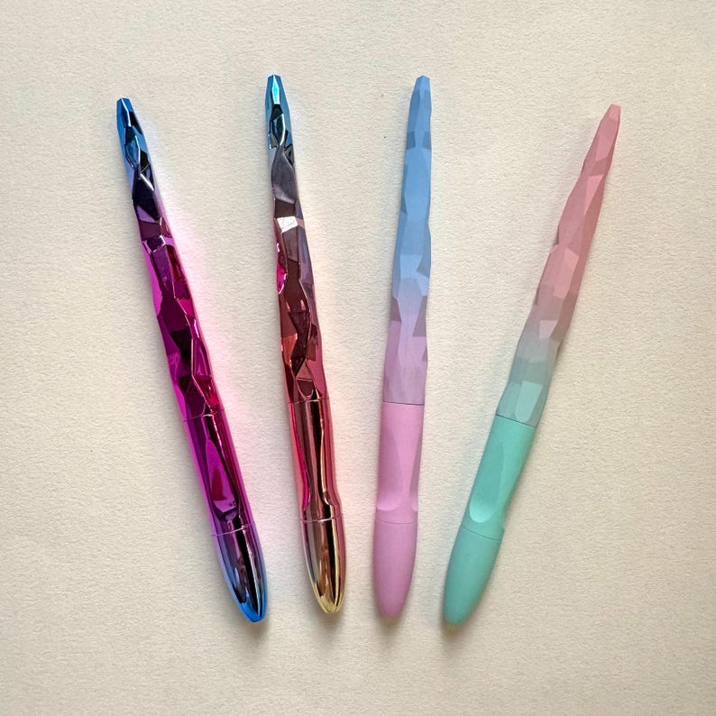 May include: Four colorful pens with faceted, jewel-like barrels. The pens transition from blue to purple, red to gold, blue to pink, and pink to green. The pens are arranged diagonally against a white background.