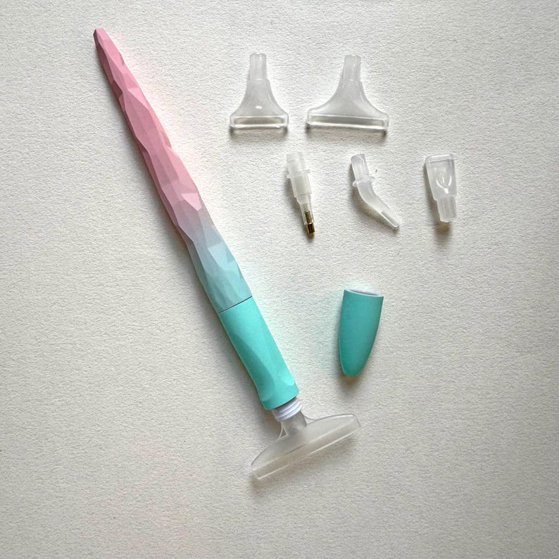 May include: A diamond painting pen with a faceted design, transitioning from pink to blue-green. Several clear plastic applicator tips and a cap are included, designed for detailed craftwork.