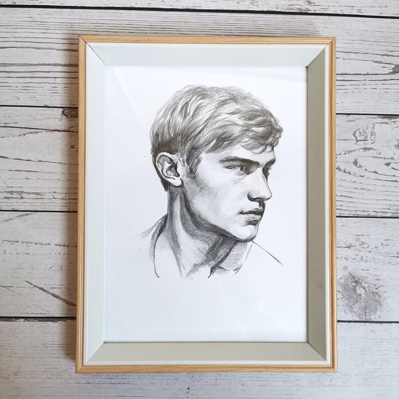 May include: A framed black and white pencil drawing of a man's face. The man has short, wavy hair and is looking to the right. The drawing is on white paper and is framed in a light wood frame.