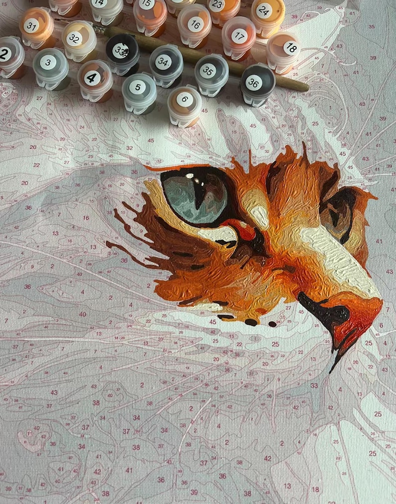 May include: A paint-by-numbers kit featuring a close-up of a cat's face. The cat's eye is a striking blue-green, and the fur is rendered in shades of orange, brown, and white. Paint pots and a brush are visible.