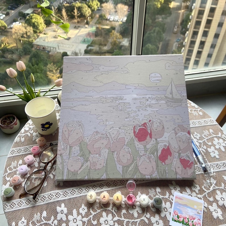 May include: A paint by numbers canvas showing a seascape with a sailboat in the distance and a field of pink tulips in the foreground. The canvas is on a table with paint pots and brushes.