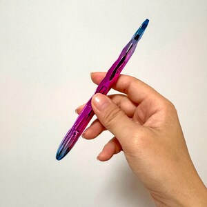 May include: A metallic pen with a vibrant color gradient, transitioning from blue to purple. The pen has a faceted design and is held in a hand against a white background. The pen's tip is a darker blue.