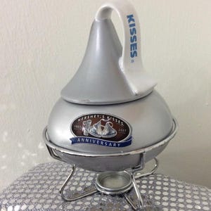 May include: Silver Hershey's Kisses 100th Anniversary hot chocolate pot with a white lid and handle. The handle has the word "KISSES" printed on it. The pot is on a silver stand with a burner.
