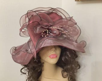 May include: A mauve-colored hat with a wide brim and a large, decorative bow. The hat features layers of sheer fabric with black trim and is adorned with small, pearl-like beads. The brim has a glittery texture.