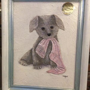 Op de afbeelding: Een ingelijst kunstwerk met een grijze puppy die een roze doek vasthoudt. De puppy heeft grote, expressieve ogen en een getextureerde vacht. Het kunstwerk staat tegen een getextureerde witte achtergrond en is ingelijst in een wit en lichtblauw frame.