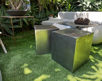 May include: Two square concrete or metal outdoor side tables in a gray color with a slightly greenish hue. The tables are sitting on a green artificial grass surface.