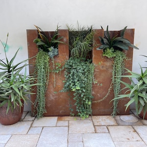May include: A rusty metal planter with three sections, each containing different types of plants. The planter is on a patio with a light grey stone floor.