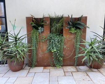 May include: A rusty metal planter with three sections, each containing different types of plants. The planter is on a patio with a light grey stone floor.