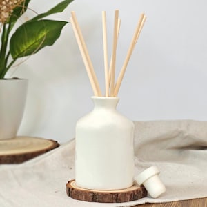 May include: A white ceramic essential oil diffuser with wooden reeds. The diffuser sits on a small, round wooden coaster. A white stopper is next to the diffuser. A plant is in the background.