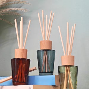 May include: Three glass reed diffusers in shades of brown, teal, and green, each with a wooden stopper and light-coloured reeds. The diffusers are arranged on a wooden shelf with books in the background.