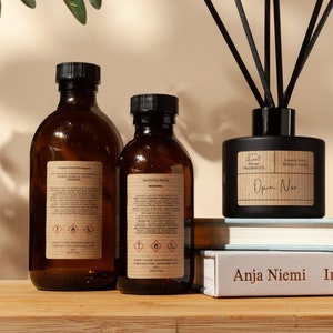 May include: Two amber glass bottles with black labels and a black glass diffuser bottle with black reeds. The bottles are on a wooden surface with a book titled "Anja Niemi In Charac" in front of them. The diffuser bottle has a label that reads "Sweet Home FRAGRANCES Luxury Reed Diffuser 330 ml Opium Noir".
