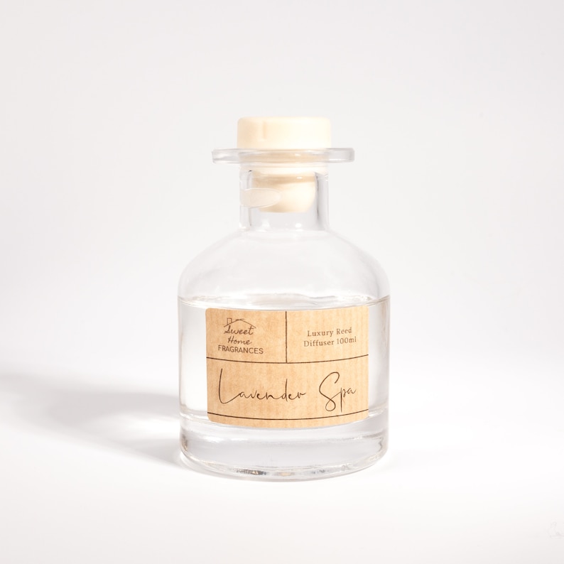 May include: A clear glass reed diffuser bottle with a light-colored wooden stopper. The bottle features a tan rectangular label with the text Sweet Home Fragrances, Luxury Reed Diffuser 100ml, and Lavender Spa printed in dark brown ink.