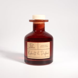 May include: A brown glass reed diffuser bottle with a white cap and a tan label. The label features the text Sweet Home Fragrances, Luxury Reed Diffuser 100ml, and Rocksalt & Driftwood. The bottle has a wide base and a narrow neck with a rim.