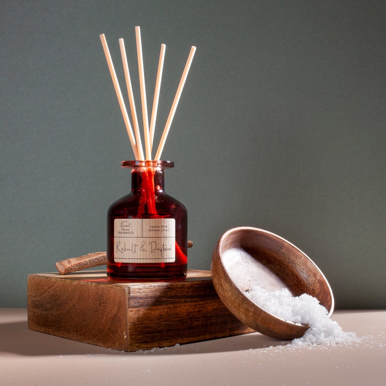 May include: A dark red glass reed diffuser bottle with five light-colored reeds, featuring a label that reads Sweet Home Fragrances and Rickult & Driftwood. Beside it sits a small wooden bowl filled with white salt crystals.