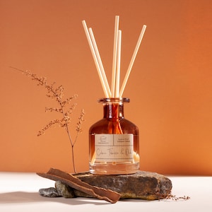 May include: A brown glass reed diffuser bottle with five light-colored reeds. The label reads Sweet Home Fragrances Luxury Reed Diffuser 100ml Cuba Tobacco & Oak. The bottle sits on a dark stone with a piece of bark and a dried plant stem.
