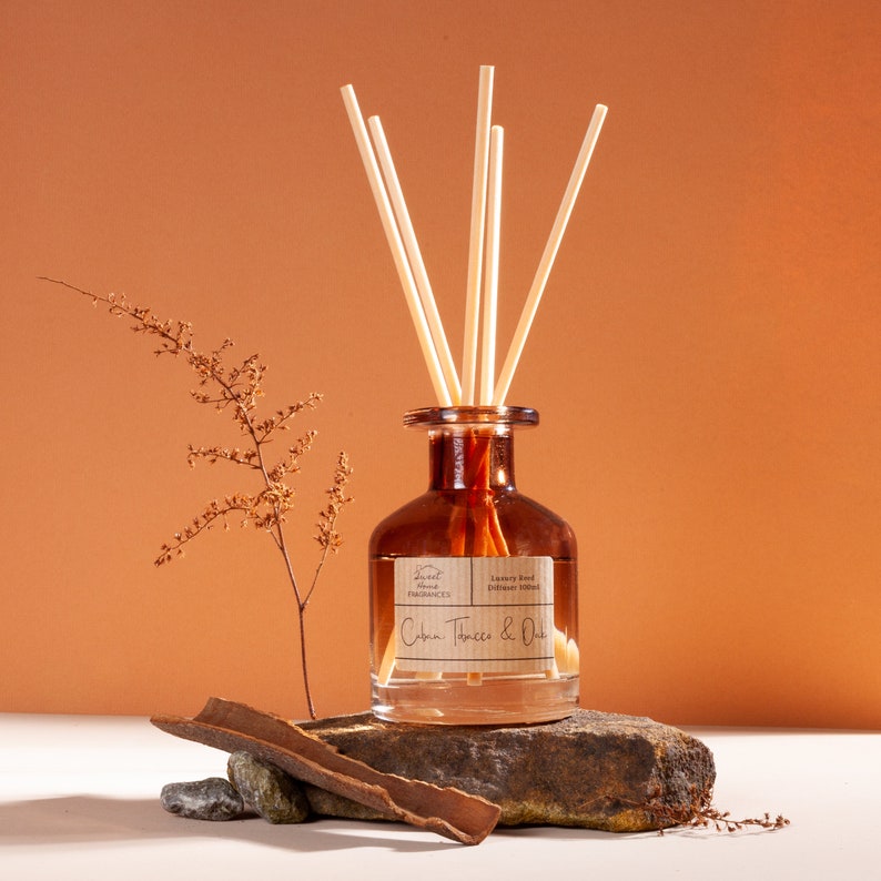 May include: A brown glass reed diffuser bottle with five light-colored reeds. The label reads Sweet Home Fragrances Luxury Reed Diffuser 100ml Cuba Tobacco & Oak. The bottle sits on a dark stone with a piece of bark and a dried plant stem.