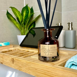 May include: A brown glass reed diffuser bottle with black reeds and a label reading Sweet Orange & Amber Luxury Reed Diffuser 100ml. The bottle sits on a bamboo tray alongside a blue and white soap bar and a light blue towel.