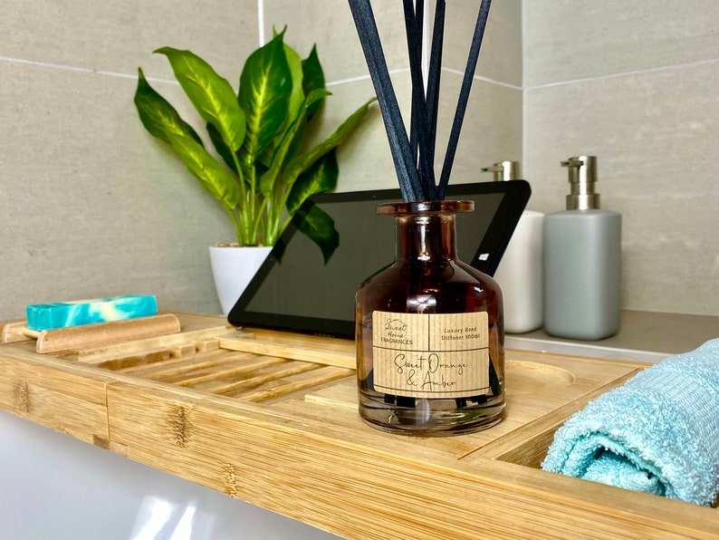 May include: A brown glass reed diffuser bottle with black reeds and a label reading Sweet Orange & Amber Luxury Reed Diffuser 100ml. The bottle sits on a bamboo tray alongside a blue and white soap bar and a light blue towel.