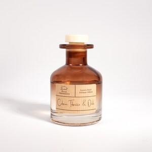 May include: A brown glass reed diffuser bottle with a white stopper. The label features the text Sweet Home Fragrances, Luxury Reed Diffuser 100ml, and Cuban Tobacco & Oak. The bottle has a wide base and a narrow neck, containing a clear liquid.