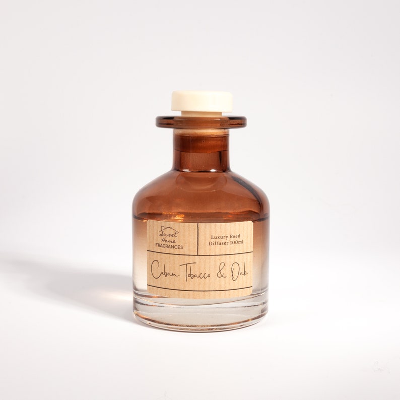 May include: A brown glass reed diffuser bottle with a white stopper. The label features the text Sweet Home Fragrances, Luxury Reed Diffuser 100ml, and Cuban Tobacco & Oak. The bottle has a wide base and a narrow neck, containing a clear liquid.