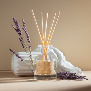 May include: A clear glass reed diffuser bottle with five light-colored wooden reeds, featuring a rectangular tan label with the text Sweet Home Fragrances, Luxury Reed Diffuser 100ml, and Lavender Spa, accompanied by sprigs of purple lavender.