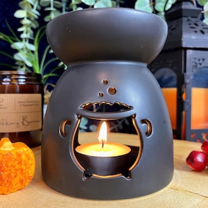 May include: Black ceramic oil burner shaped like a cauldron with a small tea light candle inside. The burner has a cauldron design with a bubbling potion effect.
