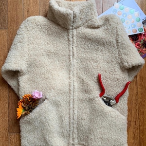 May include: Cream-colored, short-sleeved fleece jacket with a high collar and a full zipper. The jacket has two front pockets, one containing a small bouquet of flowers and the other a pair of red-handled pruning shears.