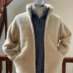 Oatmeal beige unisex wool jacket displayed on a mannequin, featuring a soft textured fabric, full zip front, high collar, and side pockets in an indoor setting