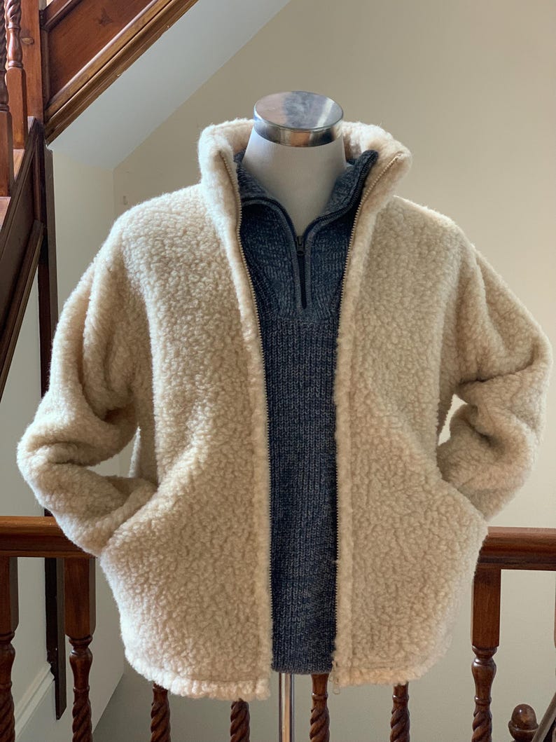 Oatmeal beige unisex wool jacket displayed on a mannequin, featuring a soft textured fabric, full zip front, high collar, and side pockets in an indoor setting